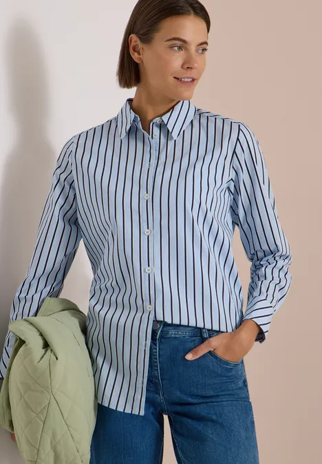 Frau in blau-weiß gestreiftem Hemd und Jeans hält eine hellgrüne Steppjacke in der Hand.