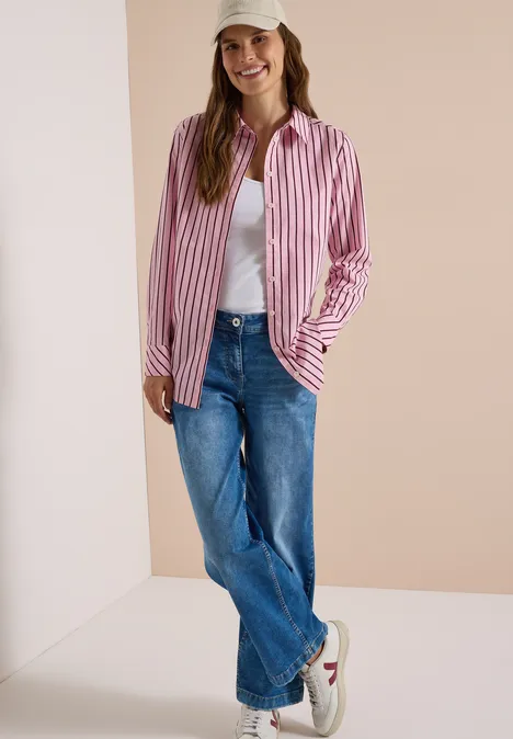 Frau trägt gestreiftes rosa-weißes Hemd, weißes Shirt, blaue Jeans und weiße Sneaker vor neutralem Hintergrund.
