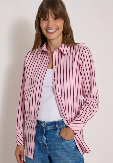 Eine Frau lächelt und trägt ein rosa-weiß gestreiftes Hemd über weißem Top und Jeans.