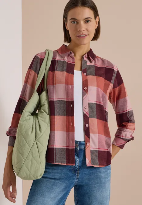 Frau in kariertem Hemd, Jeans und weißem T-Shirt trägt eine grüne gesteppte Tasche über der Schulter.