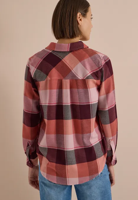 Frau im Rot- und Rosakaro-Hemd, von hinten in Jeans, vor neutralem Hintergrund.