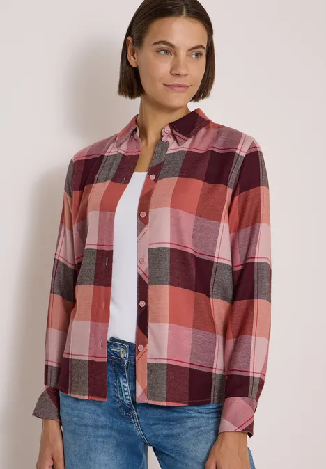 Frau in rotem und rosafarben kariertem Hemd über weißem Top und Jeans, lächelt seitlich.