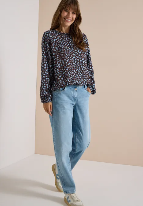 Eine Frau trägt eine gemusterte Bluse und helle Jeans, steht lässig lächelnd vor neutralem Hintergrund.