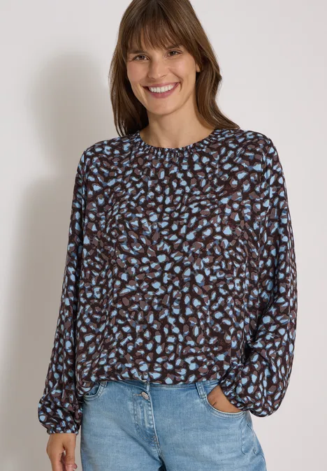 Junge Frau mit gemusterter Bluse und Jeans, sie lächelt vor hellem Hintergrund.
