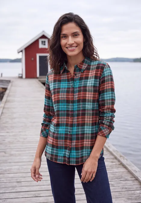 Lächelnde Frau in karierter Bluse und Jeans auf einem Holzsteg vor einem roten Bootshaus am See.