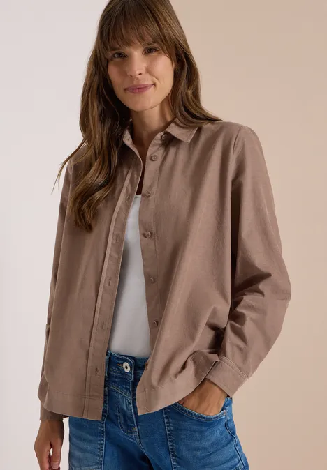 Frau in braunem Hemd, weißem Top und Jeans, mit den Händen in den Taschen, lächelnd.