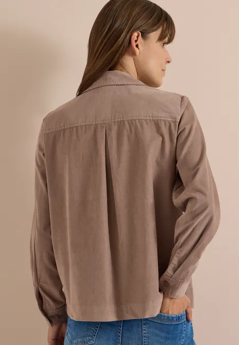 Frau in beige-brauner, locker sitzender Bluse mit langen Ärmeln, Rückenansicht vor neutralem Hintergrund.