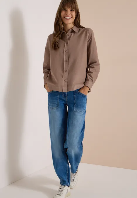 Eine lächelnde Frau in braunem Hemd und blauen Jeans vor einem hellen Hintergrund.