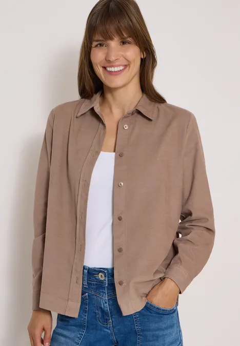 Eine lächelnde Frau trägt eine braune Hemdjacke über einem weißen Top und Jeans.