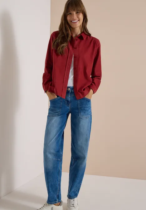 Frau mit roter Hemdjacke, weißem Shirt und blauen Jeans lehnt lächelnd an der Wand, Hände in den Taschen.