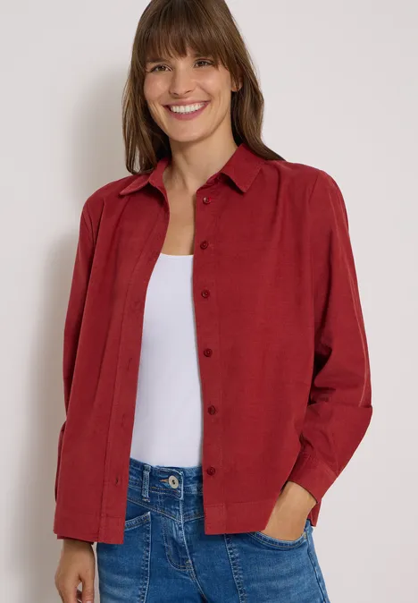 Eine lächelnde Frau trägt ein rotes Hemd über einem weißen Top und blaue Jeans.