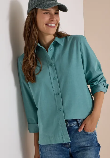 Eine Frau lehnt lächelnd an einer Wand, trägt eine grüne Bluse, Jeans und eine Schirmmütze.