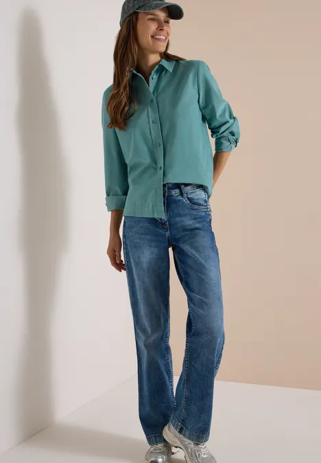 Frau in grüner Bluse, Jeans und Cap lehnt lächelnd an einer Wand.