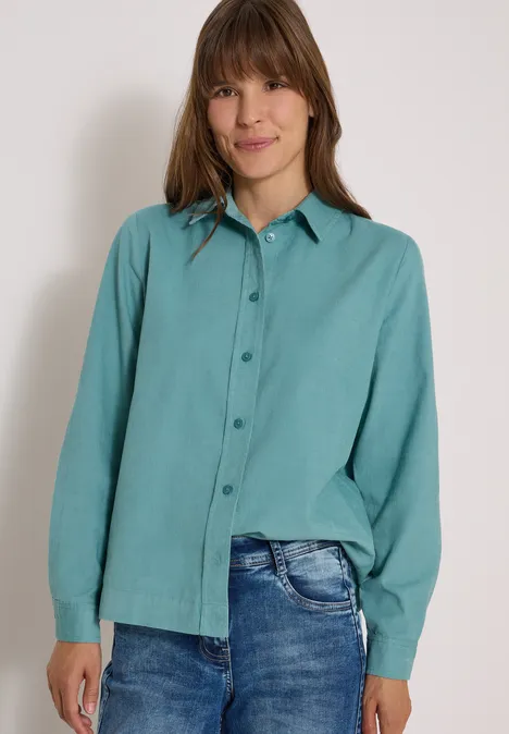 Eine Frau trägt eine türkisfarbene, langärmlige Bluse und Jeans vor neutralem Hintergrund und lächelt.