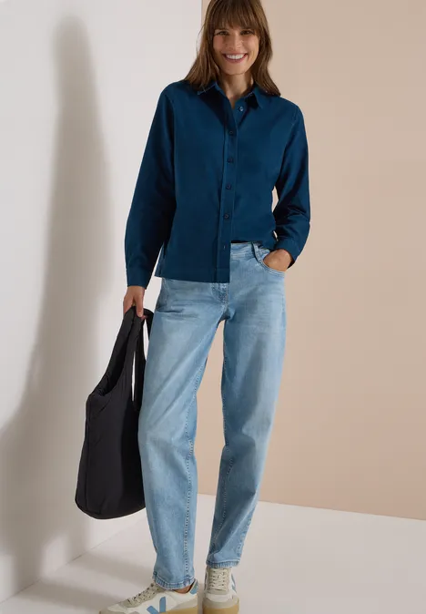 Frau in dunkelblauer Hemdbluse, hellblauen Jeans und Sneakers trägt schwarze Tasche, steht vor neutralem Hintergrund.