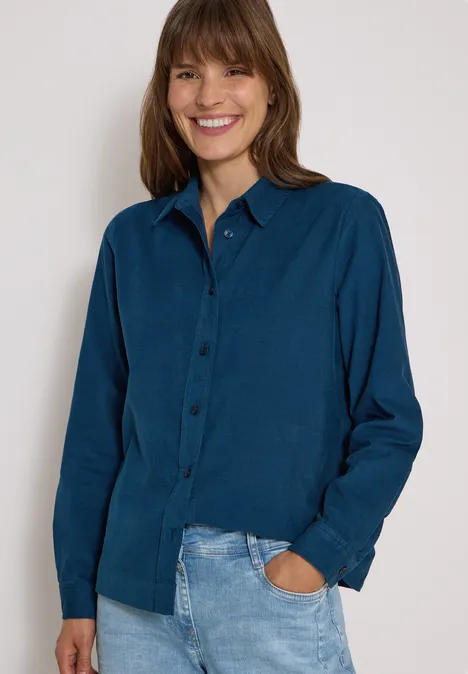 Lächelnde Frau in blauem Hemd und Jeans, mit einer Hand in der Hosentasche vor hellem Hintergrund.