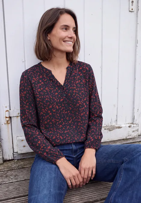 Tuniek met lange mouwen, split in de hals en print urban dark blue Een glimlachende vrouw met bruin haar draagt een roodgeprint bloes en jeans, zittend voor een witte deur.