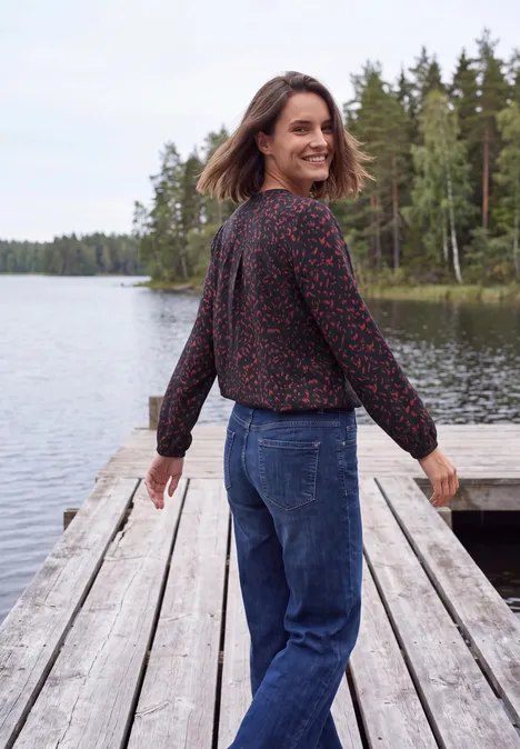Tuniek met lange mouwen, split in de hals en print urban dark blue Een vrouw met bloemenblouse en spijkerbroek staat lachend op een steiger aan een meer, bos op de achtergrond.