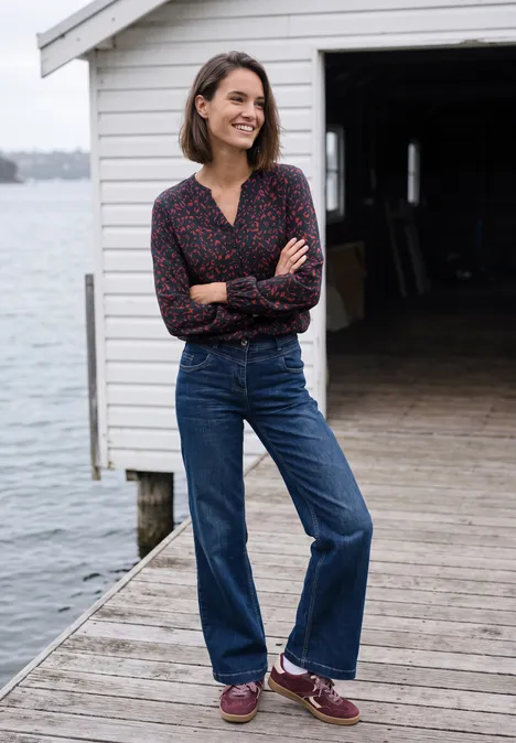 Tuniek met lange mouwen, split in de hals en print urban dark blue Een glimlachende vrouw in jeans en blouse staat met gekruiste armen op een steiger voor een wit boothuis.