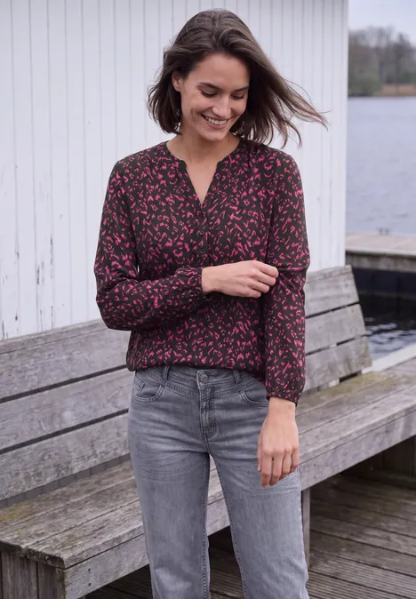 Tuniek met lange mouwen, split in de hals en print truffle brown Een jonge vrouw lacht in een bloemenblouse en grijze jeans, staand op een houten bankje aan het water.