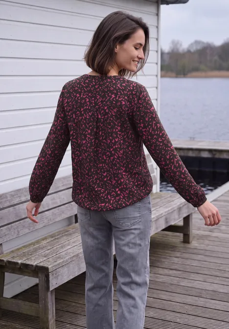 Tuniek met lange mouwen, split in de hals en print truffle brown Vrouw staat op een houten steiger bij een meer en kijkt over haar schouder, ze draagt een bloemenblouse en grijze broek.