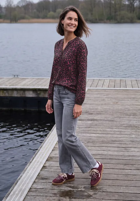 Tuniek met lange mouwen, split in de hals en print truffle brown Een vrouw in een gemusterd shirt, grijze jeans en rode sneakers staat op een steiger aan het meer.