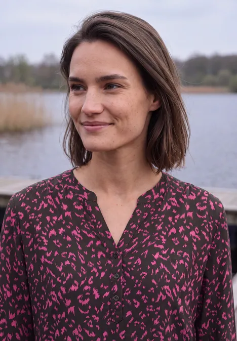 Tuniek met lange mouwen, split in de hals en print truffle brown Een glimlachende vrouw met bruin haar in een gekleurd topje bij een meer in de buitenlucht.