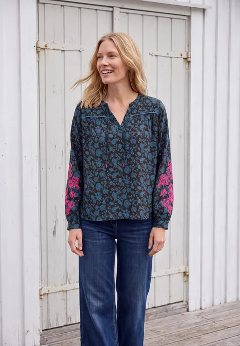Tuniek met lange mouwen, split in de hals en print truffle brown Een glimlachende vrouw in een blauw dessinblouse met roze mouwen en spijkerbroek voor een witte houten wand.