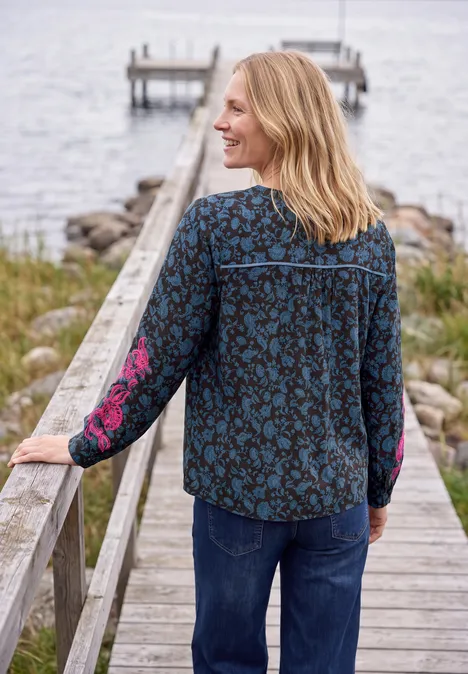 Tuniek met lange mouwen, split in de hals en print truffle brown Vrouw leunt op houten steiger bij het water, kijkt zijwaarts glimlachend, draagt gebloemde blouse en spijkerbroek.