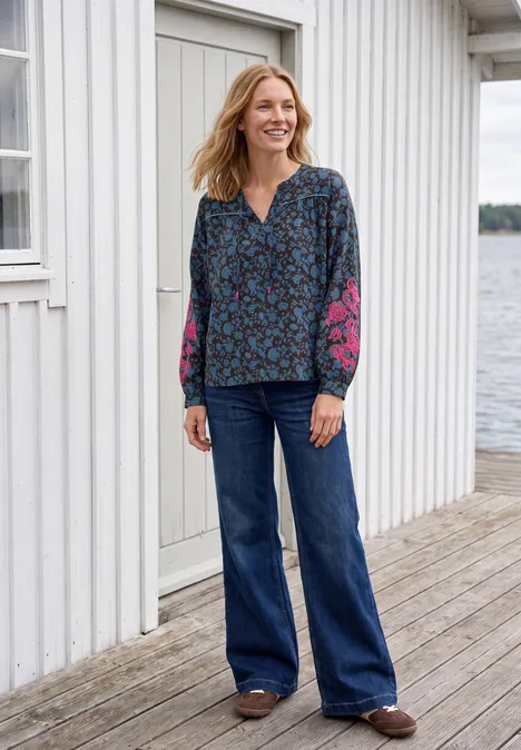 Tuniek met lange mouwen, split in de hals en print truffle brown Een glimlachende vrouw in een blauwe bloemenblouse en wijde jeans staat op een houten steiger voor een wit huis aan het water.