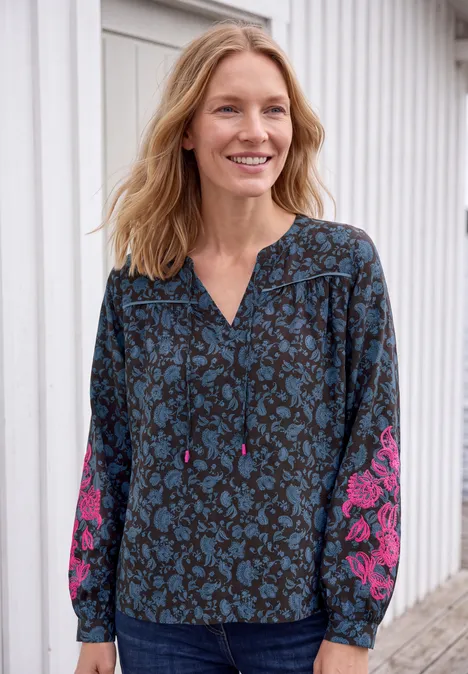 Tuniek met lange mouwen, split in de hals en print truffle brown Vrouw lacht voor witte muur, draagt blouse met blauw-roze bloemenpatroon.
