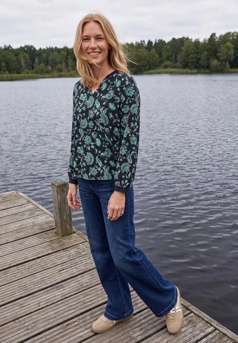 Blouse met lange mouwen, split in de hals en print urban dark blue Vrouw lacht op een steiger bij een meer, gekleed in een motiefblouse en spijkerbroek.