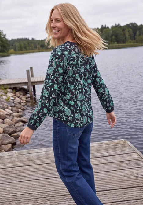 Blouse met lange mouwen, split in de hals en print urban dark blue Vrouw lacht en kijkt om op een houten steiger bij een meer, draagt groene bloemenblouse en blauwe spijkerbroek.