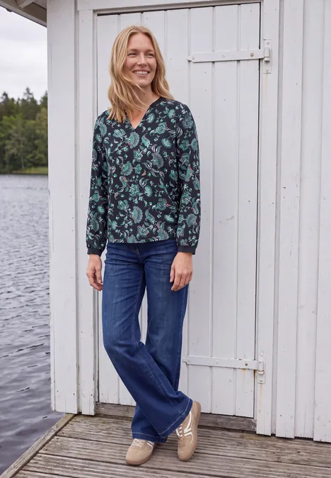Blouse met lange mouwen, split in de hals en print urban dark blue Een glimlachende vrouw in een bloemenblouse en jeans staat ontspannen op een houten steiger bij het water.