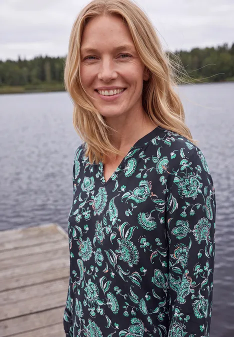 Blouse met lange mouwen, split in de hals en print urban dark blue Een lachende vrouw in een gebloemde jurk staat aan de oever van een meer.