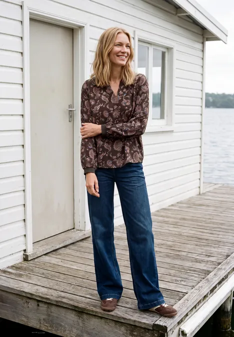 Lächelnde Frau in gemusterter Bluse und Jeans auf einem Steghaus am Wasser.