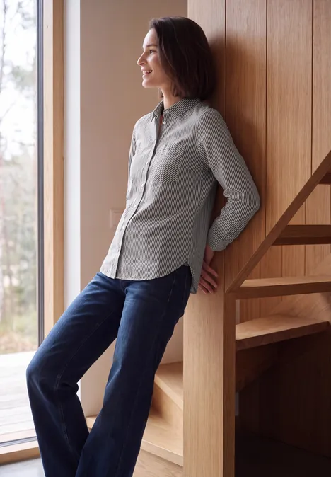 Seersucker Hemdbluse mit Streifen truffle brown Eine Frau lehnt entspannt am Holzgeländer neben einem Fenster, lässig in Hemd und Jeans.