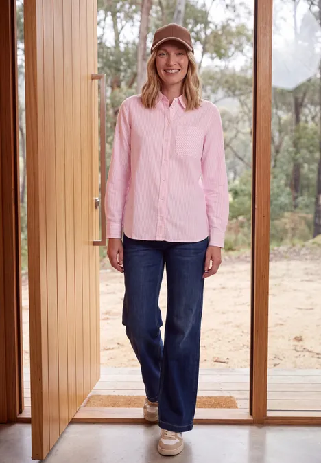 Overhemd met lange mouwen en strepen mellow rose Een glimlachende vrouw in spijkerbroek, roze blouse en pet staat in een openstaande deur voor een bos.