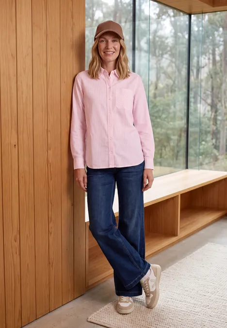 Overhemd met lange mouwen en strepen mellow rose Vrouw in roze blouse, blauwe jeans en pet leunt glimlachend tegen een houten wand bij grote ramen.