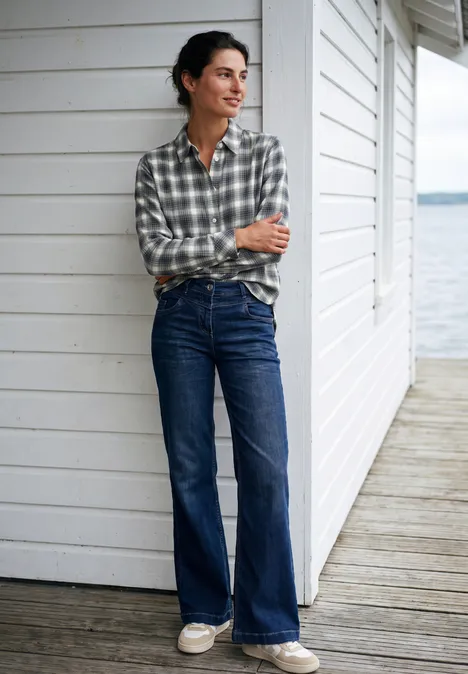 Flanellen blouse met lange mouwen en ruitjespatroon vanilla white Vrouw in geruit overhemd en wijde jeans staat met gekruiste armen voor een wit houten huis en kijkt opzij.