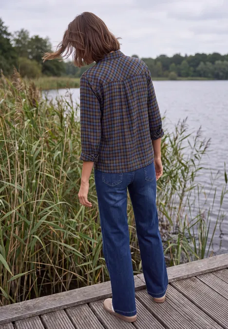 Persoon in geruit overhemd en jeans op een houten steiger bij een meer, omgeven door riet en bomen.