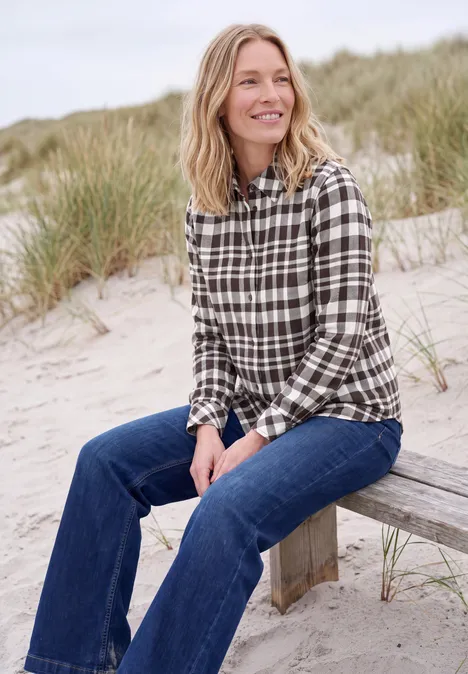 Langarm Flanell Bluse mit Karomuster truffle brown Eine lächelnde Frau sitzt auf einer Holzbank am Strand mit kariertem Hemd und Jeans.