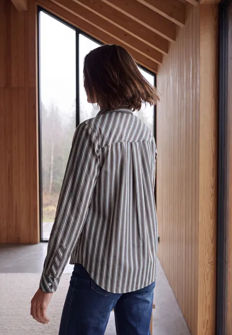 Langarm Hemdbluse mit Streifen truffle brown Eine Frau trägt ein gestreiftes Hemd und Jeans, steht seitlich in einem Holzraum und blickt aus dem Fenster.