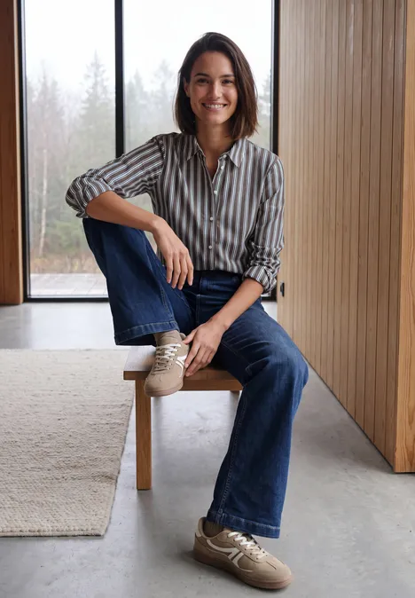 Langarm Hemdbluse mit Streifen truffle brown Eine Frau sitzt lächelnd auf einer Holzbank vor Fenster, trägt Streifenbluse, Jeans und Sneaker.