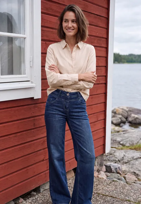 Langarm Hemdbluse aus Cord in Unifarbe cashmere beige Frau in Jeans und hellem Hemd vor rotem Holzhaus am See, verschränkt die Arme und lächelt.