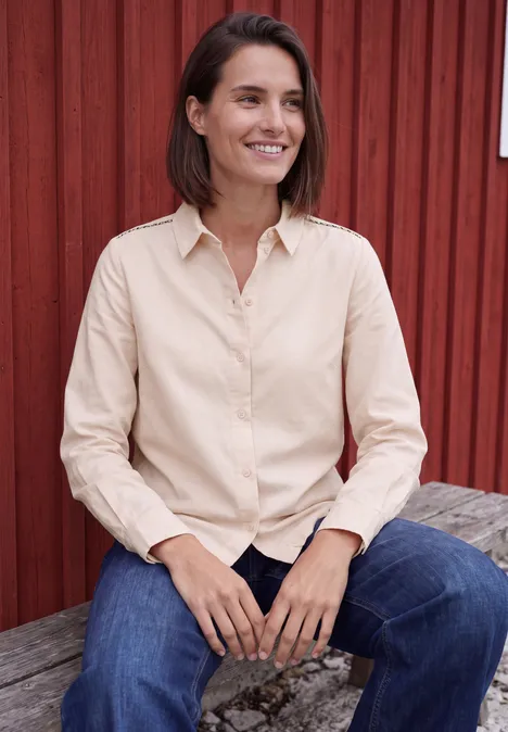 Langarm Hemdbluse aus Cord in Unifarbe cashmere beige Eine lächelnde Frau sitzt auf einer Holzbank vor einer roten Wand, trägt beiges Hemd und blaue Jeans.