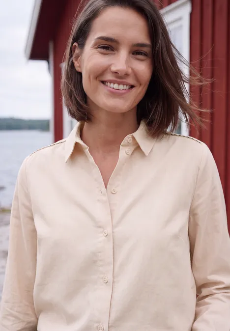 Langarm Hemdbluse aus Cord in Unifarbe cashmere beige Eine lächelnde Frau im hellen Hemd vor Wasser und rotem Holzhaus.