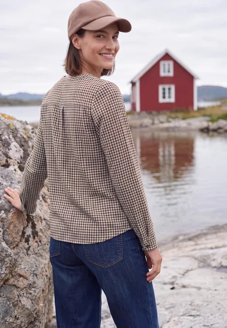 Tuniek met lange mouwen, split in de hals en print mocha brown Een vrouw in geruit shirt, spijkerbroek en pet lacht aan de oever, met een rood huis op de achtergrond.