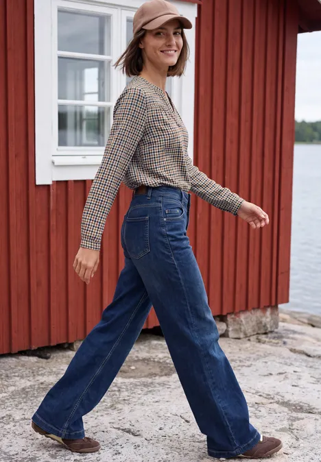 Tuniek met lange mouwen, split in de hals en print mocha brown Vrouw met pet, geruit overhemd en wijde jeans, lopend voor een rood houten huis aan het water.