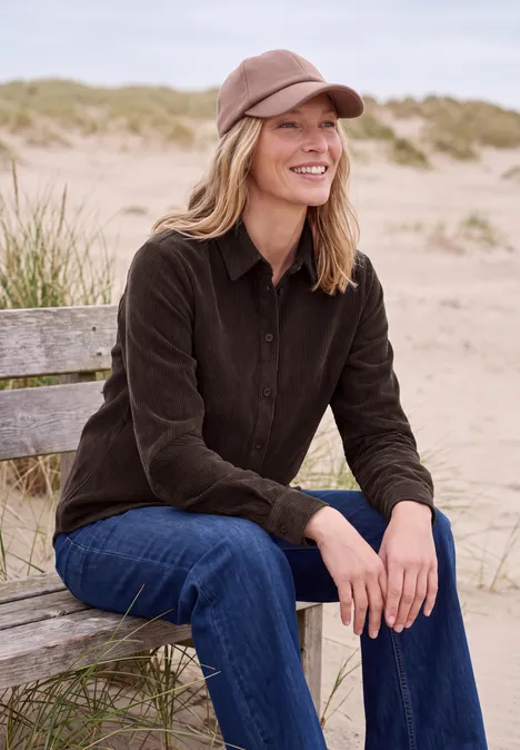Langarm Hemdbluse aus Cord in Unifarbe truffle brown Eine lächelnde Frau in brauner Jacke und Cap sitzt entspannt auf einer Bank am Strand mit Dünen im Hintergrund.